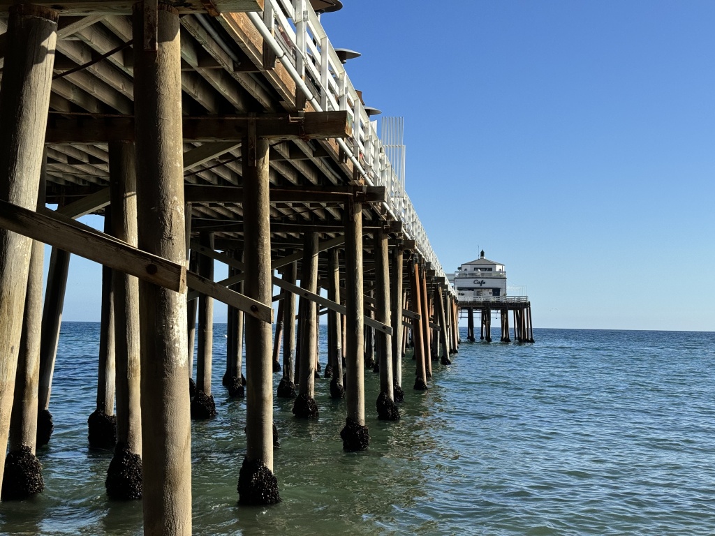 A photo from the underside of a pier with the end is sight and the ocean surrounding it on all sides.