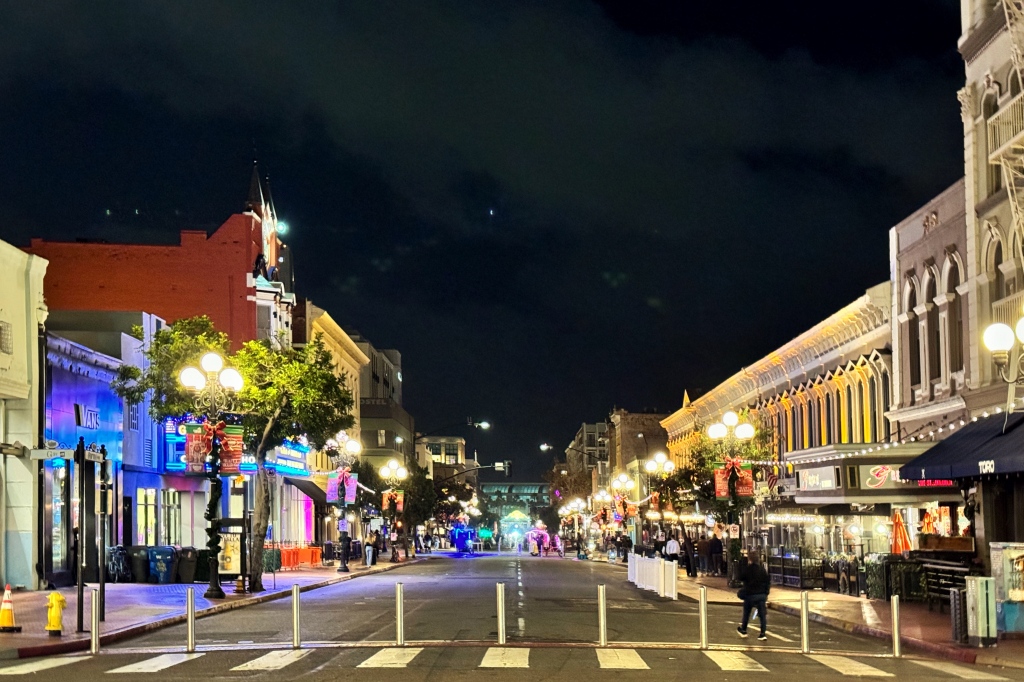 A street lined with bollards, shops on either end and bright lights on each end.